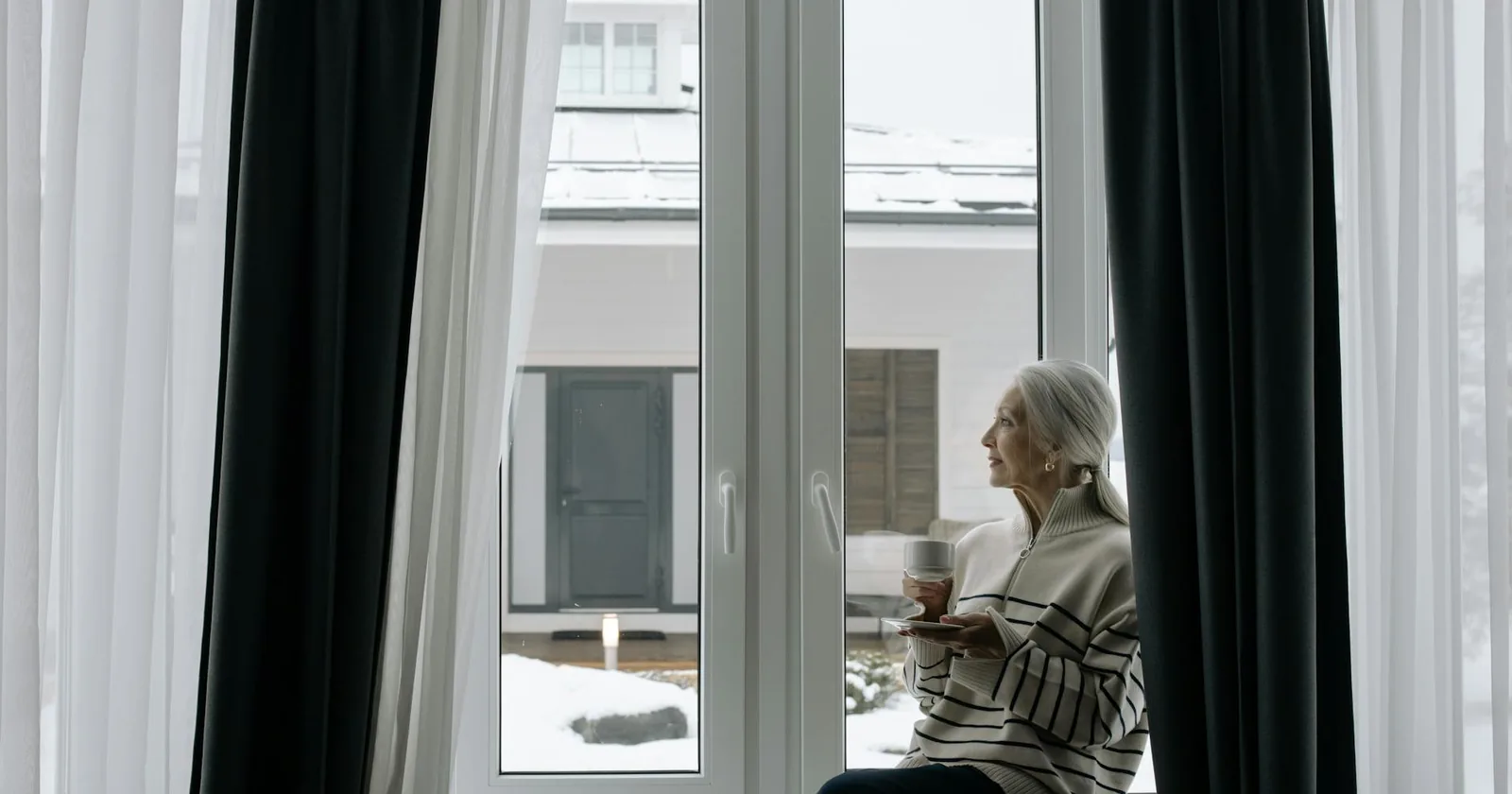 Older woman sitting alone by a sunlit window at home, reflecting on the choice between staying put and moving to a senior community.