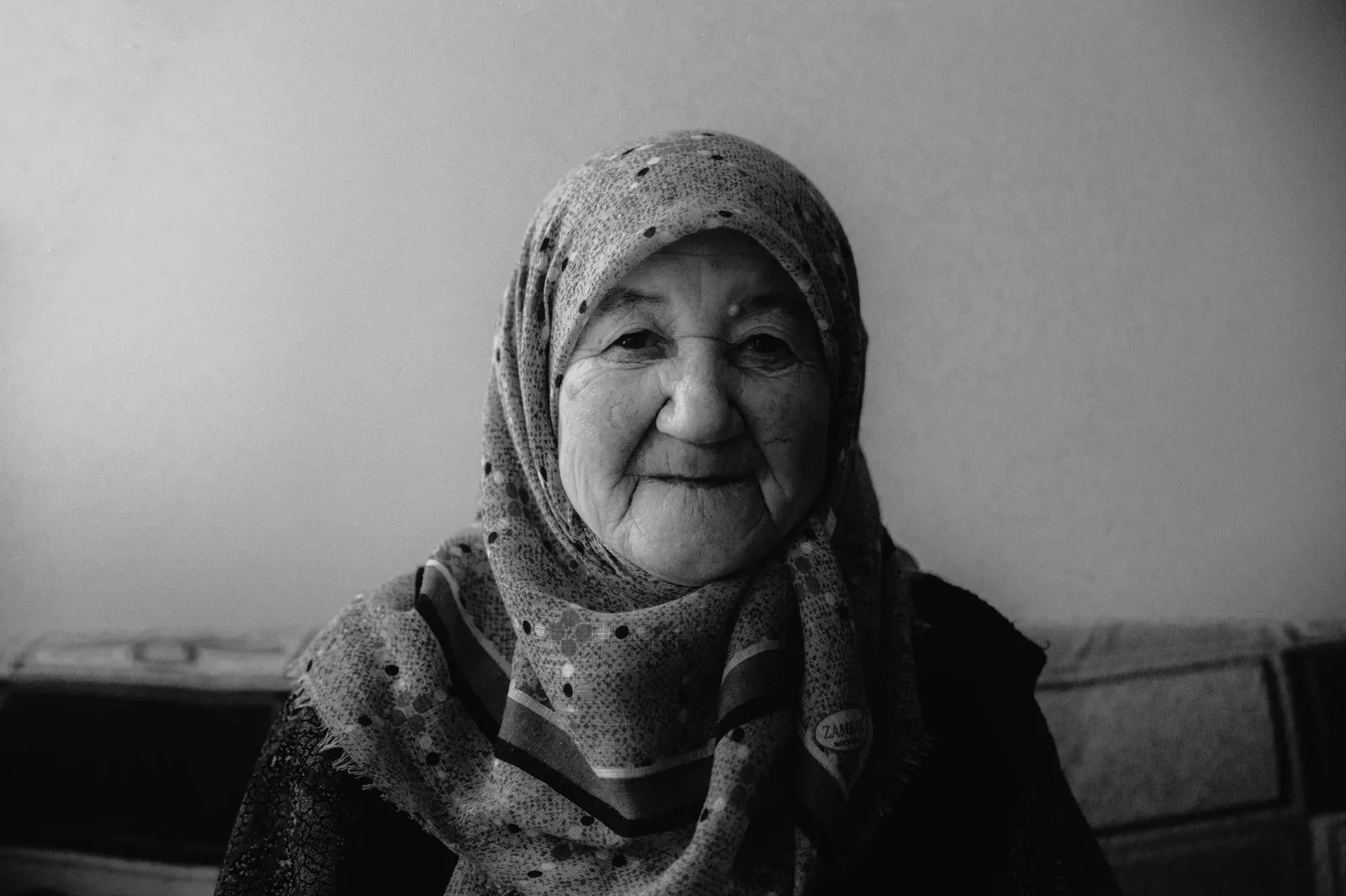 Black and white portrait of an elderly woman smiling warmly, wearing a hijab indoors.
