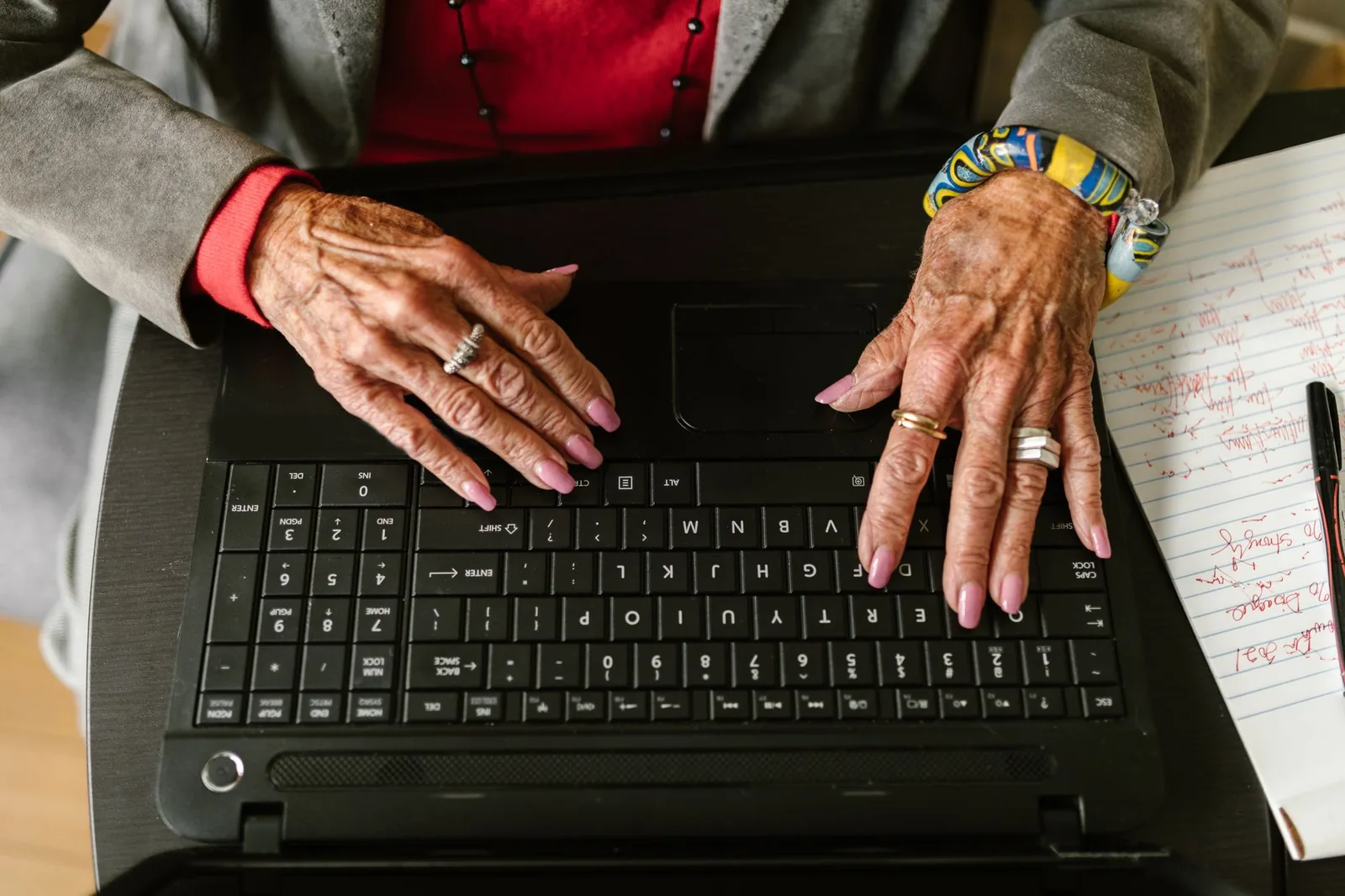 Close-up of an older woman's hands typing on a laptop keyboard beside a handwritten notebook and pen.