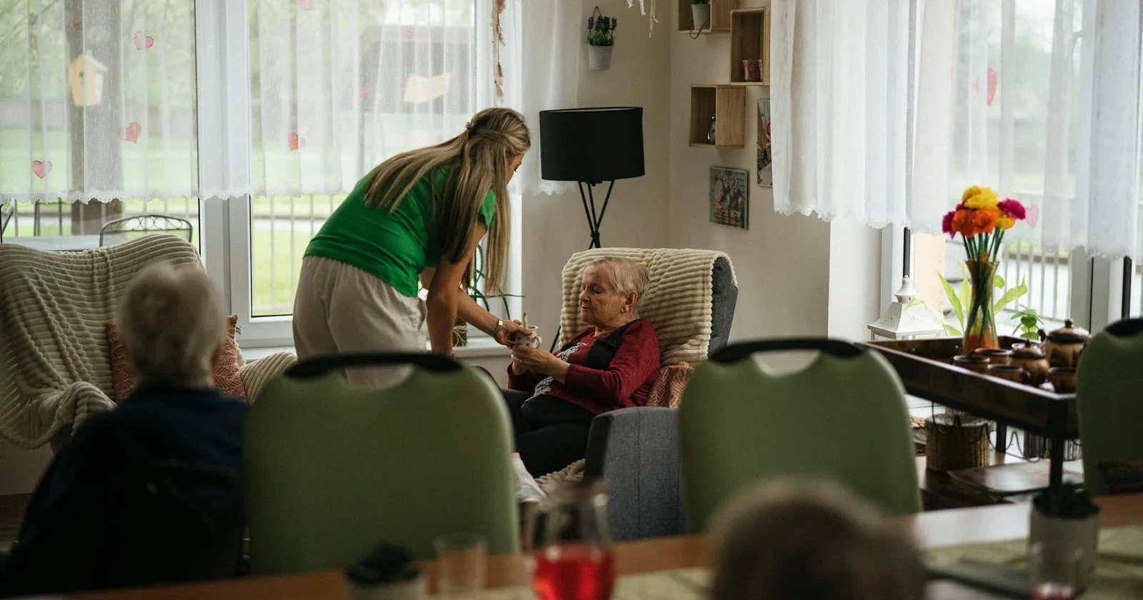 Caregiver assisting elderly women with tea in a cozy senior living community setting.