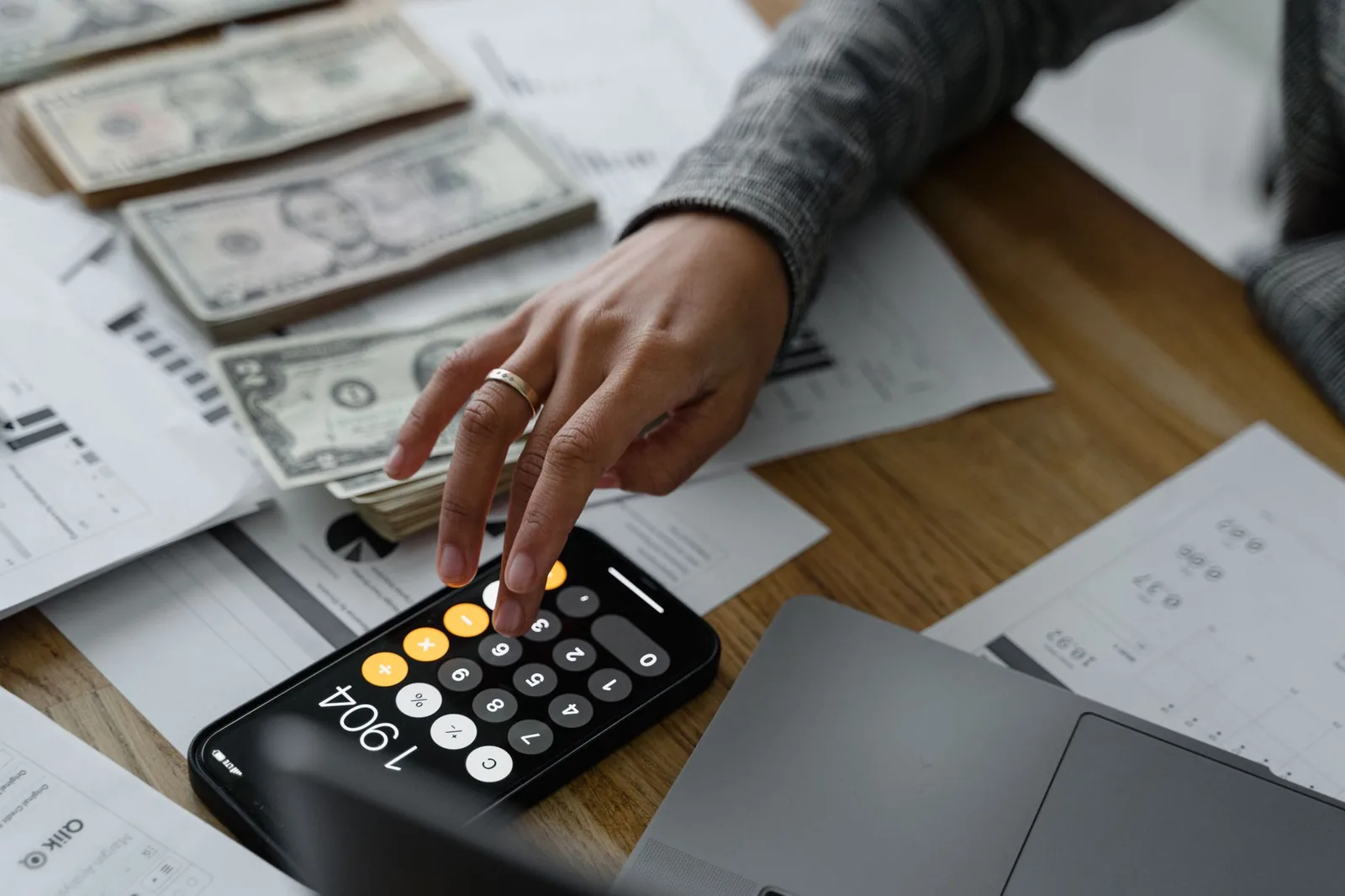 A hand working a calculator beside stacks of cash and printed financial statements on a wooden table.