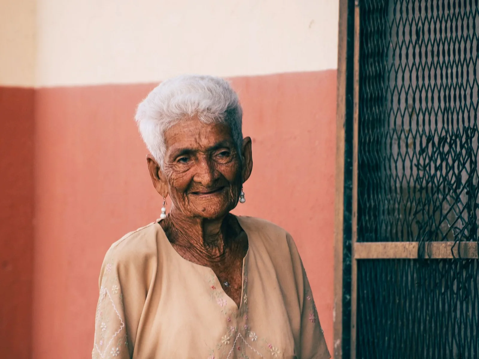An elderly woman with white hair smiles warmly in front of a colorful wall.