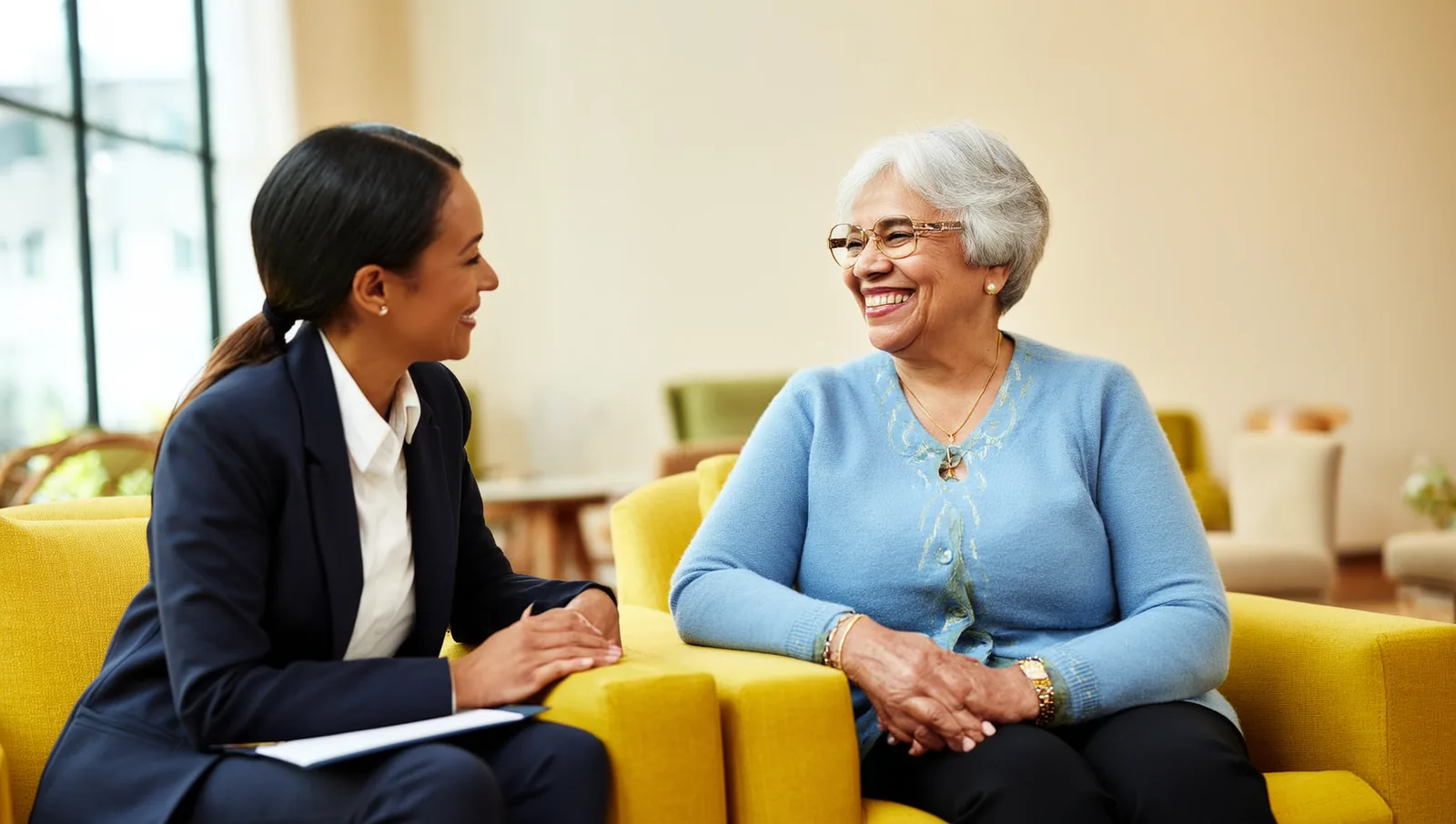 A resident advocate sitting and listening attentively to an elderly woman in a bright assisted living lounge.