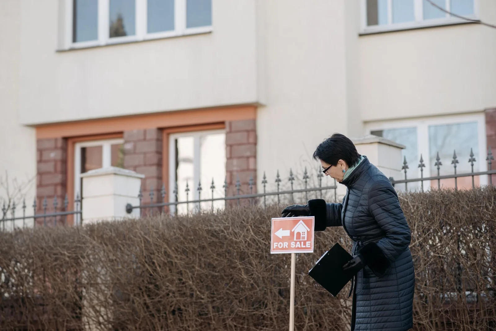 A real estate agent in a dark coat setting a For Sale sign into a hedge in front of a house.