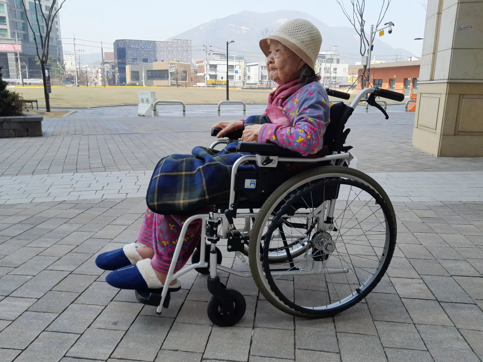 An older woman seated in a wheelchair on a paved plaza outside, with the town and hills behind her.
